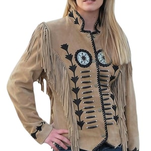 Könnte beinhalten: Hellbraune Wildlederjacke mit Fransen an den Ärmeln und am Rücken. Die Jacke hat schwarze und weiße Perlenverzierungen und dekorative Nähte. Sie hat eine Knopfleiste und einen hohen Kragen. Die Ärmel sind lang, getragen mit blauen Jeans.