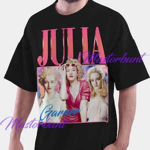 以下が含まれることがあります： ピンクのグラデーションで「JULIA」と書かれた黒いTシャツ。長方形の画像の上にあります。画像には女性の3つの肖像画が写っています。「Garner Masterbunt」というテキストが筆記体で書かれています。