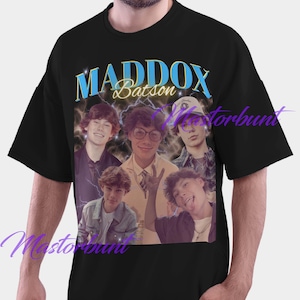 May include: Black t-shirt featuring a graphic of five young men with the text "MADDOX Batson" in blue and gold. The design has a vintage, retro style with a lightning bolt effect.