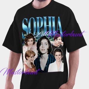 May include: Black t-shirt with a design featuring multiple images of Sophia Lillis, with the name "Sophia Lillis" in a blue, stylized font. The design includes several photos of the actress.
