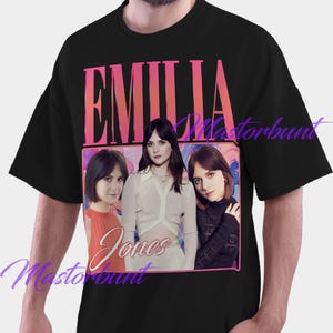 May include: Black t-shirt featuring a graphic of Emilia Jones with the name "Emilia" in pink and orange block letters above the image. The name "Jones" is in cursive below the image. The image is framed in a pink border.
