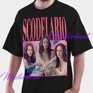 Op de afbeelding: Zwart T-shirt met een roze en oranje afbeelding van de naam "Scodelario" boven een foto van drie vrouwen. De foto is omlijst in roze en geplaatst tegen een kleurrijke achtergrond.