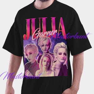以下が含まれることがあります： 黒いTシャツ。ジュリア・ガーナーのグラフィックと「JULIA」という名前がピンクとゴールドの文字で描かれています。グラフィックには、紫と青の銀河の背景に複数の女優の画像が含まれています。このシャツはカジュアルなスタイルです。