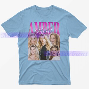 May include: Light blue t-shirt featuring a collage of images of Amber Heard, with the name "AMBER Heard" in pink and white text. The shirt is a crew neck style.