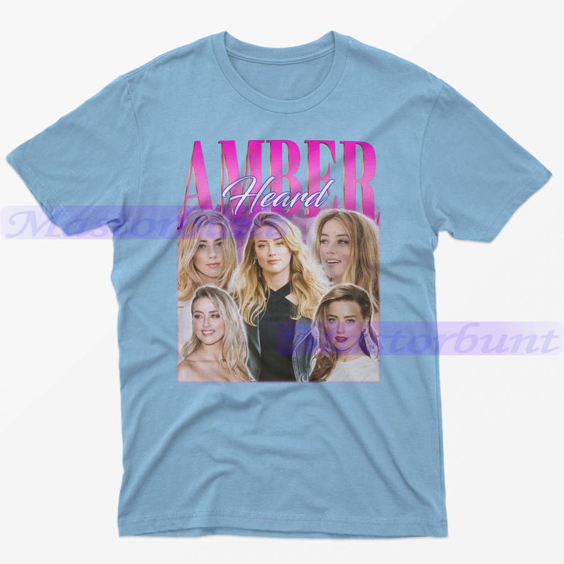 May include: Light blue t-shirt featuring a collage of images of Amber Heard, with the name "AMBER Heard" in pink and white text. The shirt is a crew neck style.