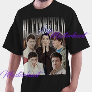 Puede incluir: Camiseta negra con un collage de imágenes de Asa Butterfield. El nombre "BUTTERFIELD" está en plata sobre "Asa" en cursiva. Un paisaje urbano forma el fondo del diseño.