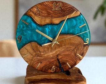 May include: A circular desk clock crafted from natural olive wood with a vibrant turquoise resin river running through the center. The clock face features gold-colored hands and simple gold-colored hour markers, resting on a matching wood base.