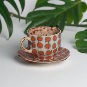 May include: A ceramic teacup and saucer set with a cream-coloured base and a pattern of orange circles connected by blue lines. The cup has a handle and sits on a matching saucer. Green leaves are in the background.