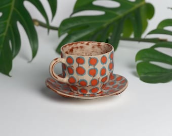 May include: A ceramic teacup and saucer set with a cream-coloured base and a pattern of orange circles connected by blue lines. The cup has a handle and sits on a matching saucer. Green leaves are in the background.