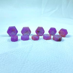 May include: A collection of eight purple gemstones, cut into hexagonal shapes, arranged in a row on a white surface.