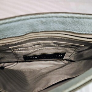 May include: Open view of a gray suede handbag interior. The bag has multiple compartments and a black label. The interior lining is a light gray fabric. The exterior of the bag is a muted gray color.