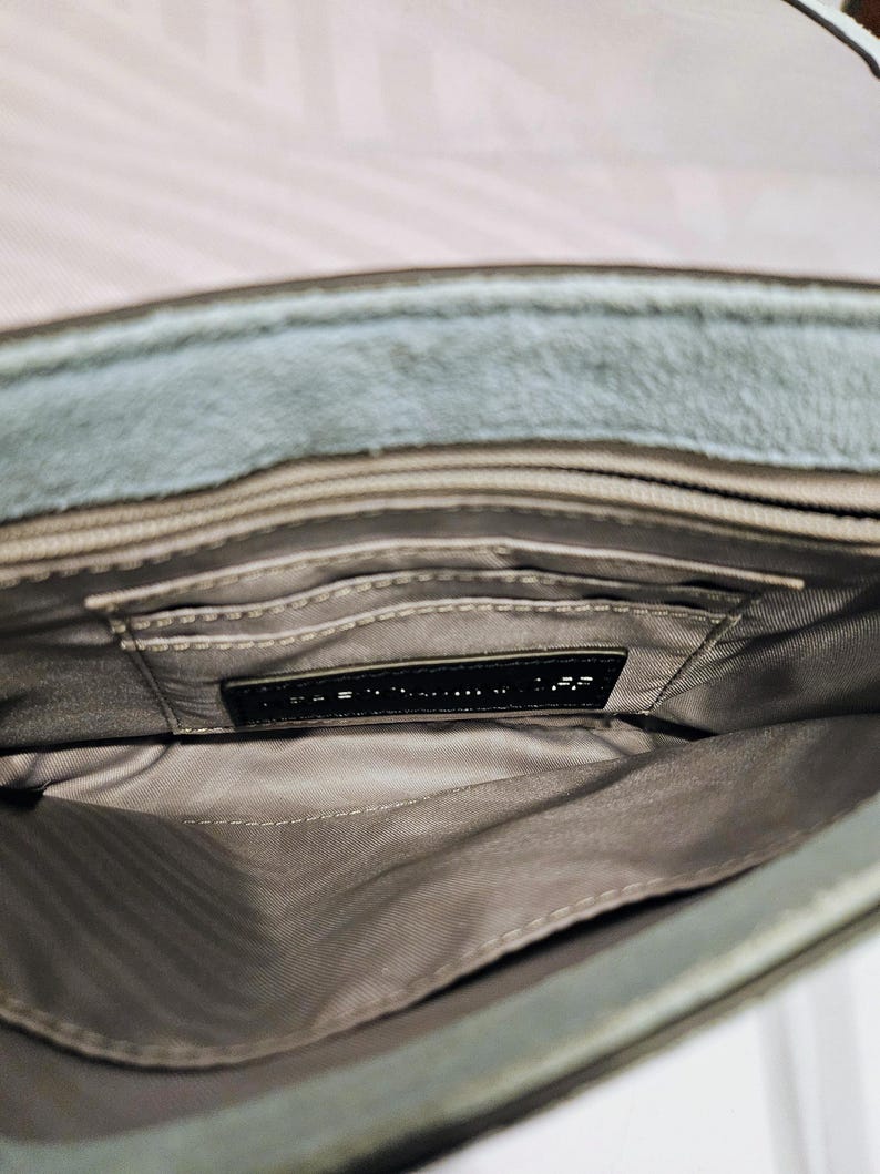 May include: Open view of a gray suede handbag interior. The bag has multiple compartments and a black label. The interior lining is a light gray fabric. The exterior of the bag is a muted gray color.