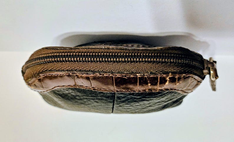 May include: A small, brown leather coin purse with a zipper closure. The purse has a dark brown zipper and a lighter brown leather accent at the top. The bottom portion of the purse is a darker shade of brown.