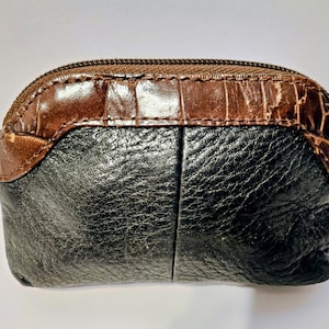 May include: A small, oval-shaped coin purse with a brown, textured top and a black, textured bottom. The purse has a zipper closure and a small, metal zipper pull. The top section has a crocodile skin pattern.