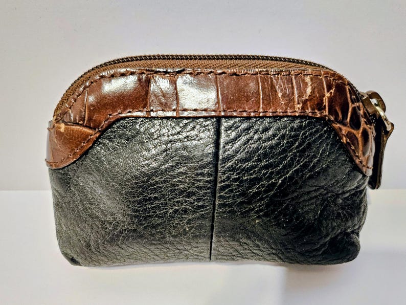 May include: A small, oval-shaped coin purse with a brown, textured top and a black, textured bottom. The purse has a zipper closure and a small, metal zipper pull. The top section has a crocodile skin pattern.