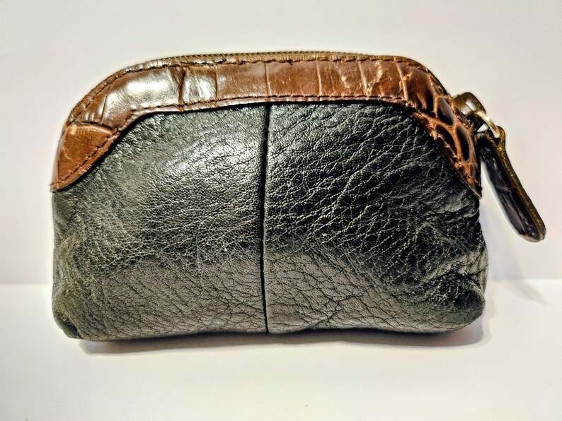 May include: A small, textured coin purse with a brown, crocodile-embossed top and a black, leather-like body. The purse has a zipper closure with a brown pull tab. The overall shape is rounded and compact.