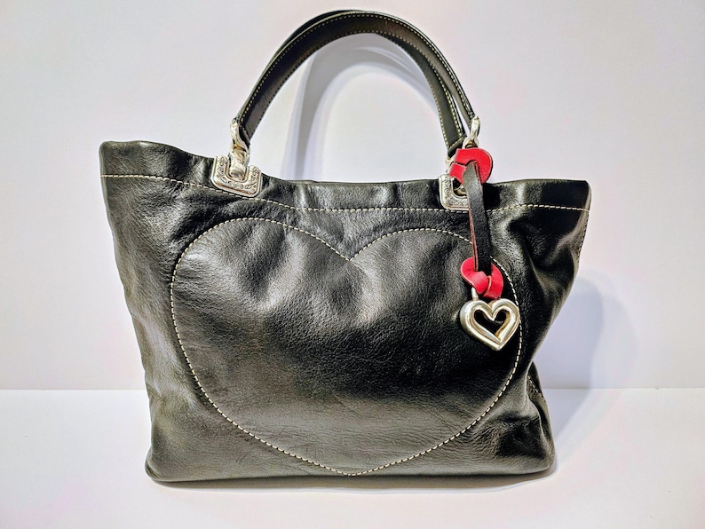 Puede incluir: Bolso tote de cuero negro con un dise&ntilde;o en forma de coraz&oacute;n cosido en la parte delantera. El bolso presenta fijaciones de asa de metal plateado y un charm de coraz&oacute;n rojo y plateado. El bolso tiene un asa superior.