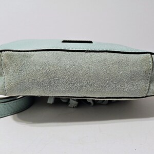 May include: A light blue crossbody bag with a suede bottom and a leather strap. The bag has a rectangular shape with a black trim and a small black accent on the top. The bottom of the bag has fringe.