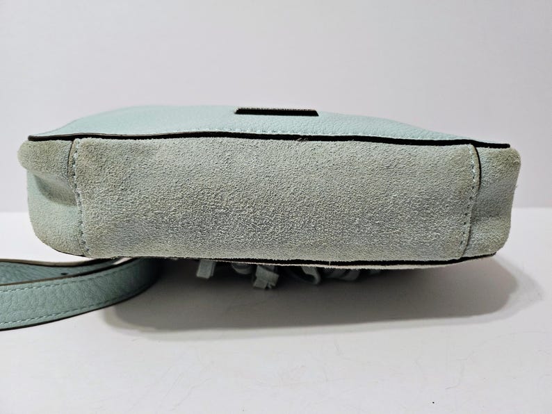 May include: A light blue crossbody bag with a suede bottom and a leather strap. The bag has a rectangular shape with a black trim and a small black accent on the top. The bottom of the bag has fringe.