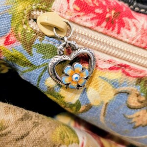 Puede incluir: Primer plano de un bolso con estampado floral con una cremallera color crema y un dije en forma de coraz&oacute;n. El dije presenta una flor azul y naranja con un peque&ntilde;o centro de cristal. La tela del bolso tiene un dise&ntilde;o floral azul, rojo y amarillo.