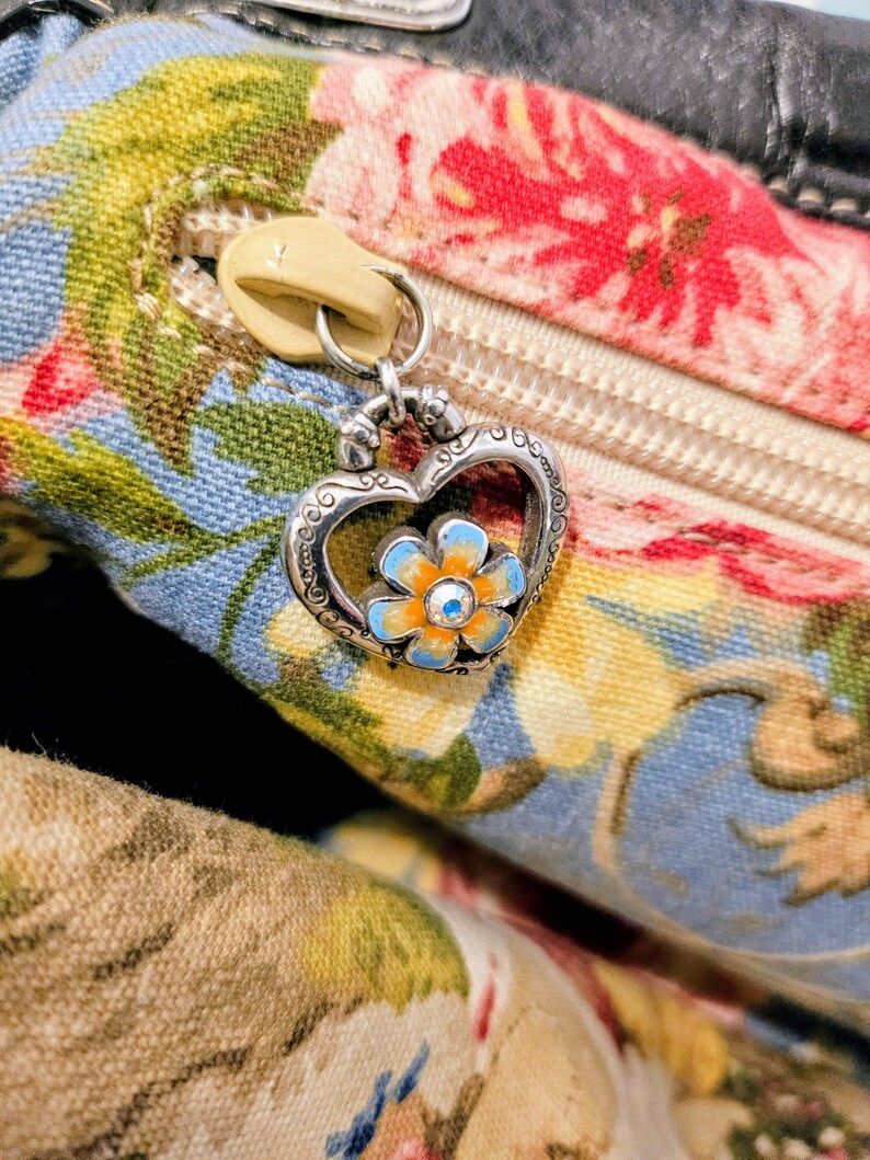 Puede incluir: Primer plano de un bolso con estampado floral con una cremallera color crema y un dije en forma de coraz&oacute;n. El dije presenta una flor azul y naranja con un peque&ntilde;o centro de cristal. La tela del bolso tiene un dise&ntilde;o floral azul, rojo y amarillo.