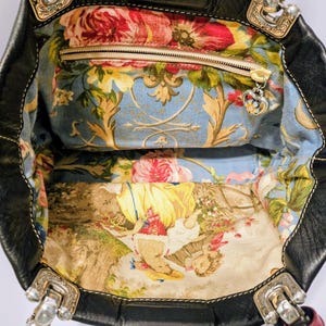 Puede incluir: Bolso tote de cuero negro con forro de tela floral. El interior cuenta con un bolsillo con cremallera y un charm decorativo. Los herrajes plateados acent&uacute;an el exterior del bolso, y un charm en forma de coraz&oacute;n cuelga del asa.