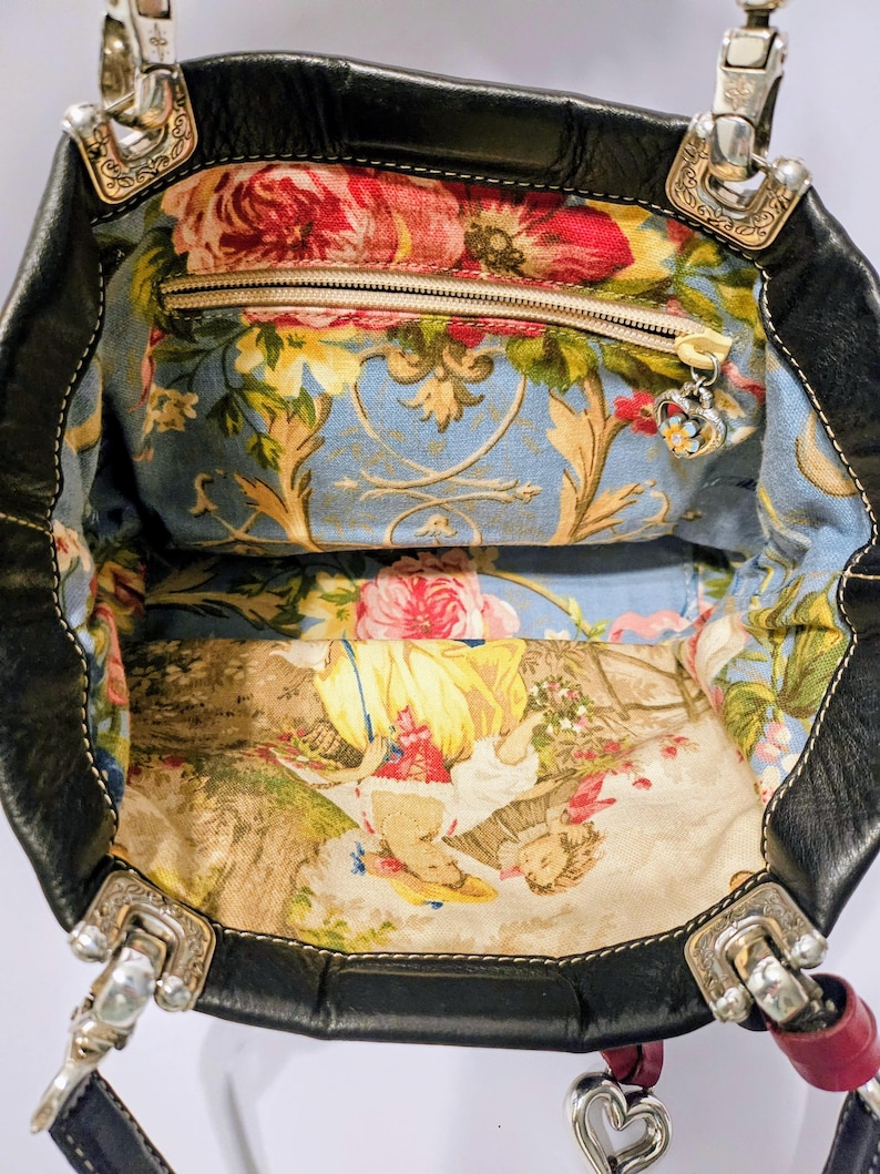 Puede incluir: Bolso tote de cuero negro con forro de tela floral. El interior cuenta con un bolsillo con cremallera y un charm decorativo. Los herrajes plateados acent&uacute;an el exterior del bolso, y un charm en forma de coraz&oacute;n cuelga del asa.