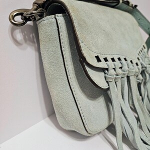 May include: A light blue suede crossbody bag with a flap closure and fringe detailing. The bag features a silver-tone clasp and a matching adjustable strap. The bag's design includes decorative cutouts and a stitched border.