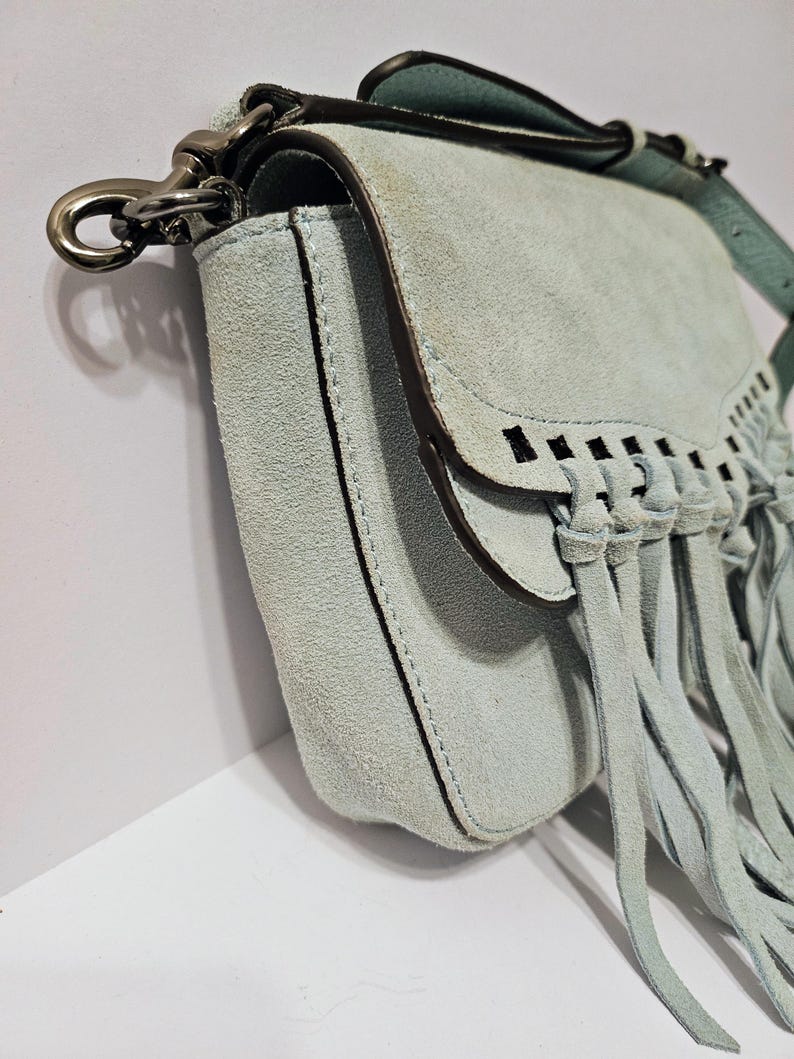 May include: A light blue suede crossbody bag with a flap closure and fringe detailing. The bag features a silver-tone clasp and a matching adjustable strap. The bag's design includes decorative cutouts and a stitched border.