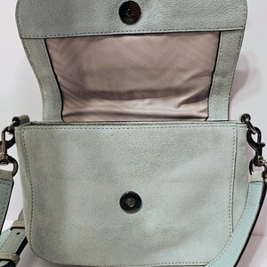 May include: A light blue suede crossbody bag with a flap closure and a long adjustable strap. The bag features a magnetic snap closure and a light brown interior. The top of the bag has a fringe detail. The bag's dimensions are not visible.