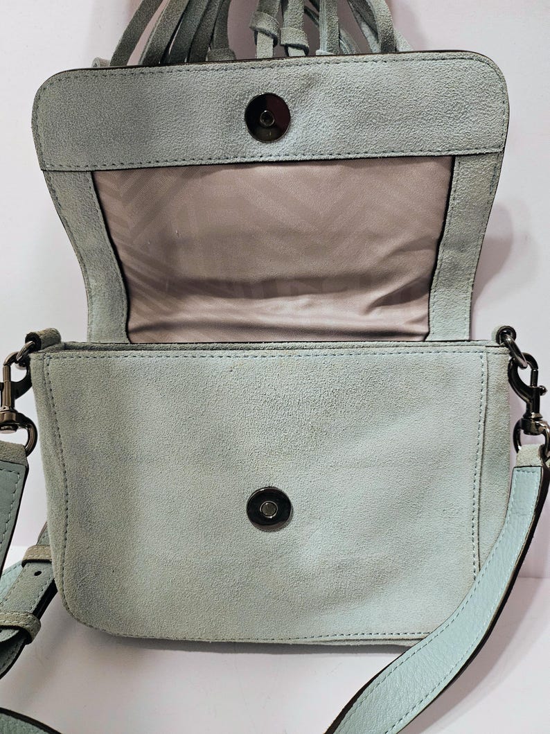 May include: A light blue suede crossbody bag with a flap closure and a long adjustable strap. The bag features a magnetic snap closure and a light brown interior. The top of the bag has a fringe detail. The bag's dimensions are not visible.