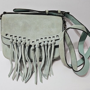 May include: A light blue suede crossbody bag with a flap closure and fringe detailing. The bag features a long, adjustable strap and a short handle. The bag's dimensions are approximately 8 inches wide, 6 inches high, and 3 inches deep.