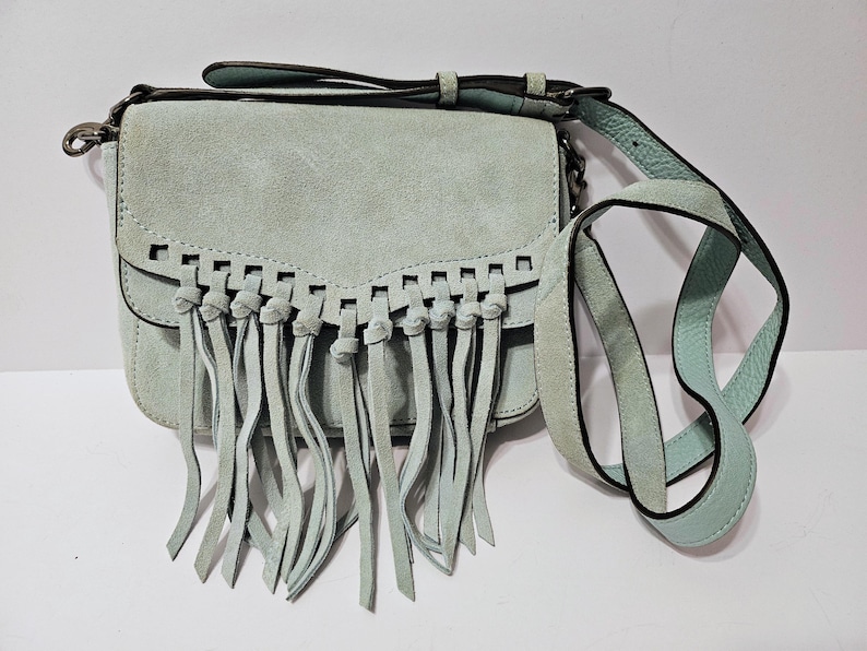 May include: A light blue suede crossbody bag with a flap closure and fringe detailing. The bag features a long, adjustable strap and a short handle. The bag's dimensions are approximately 8 inches wide, 6 inches high, and 3 inches deep.