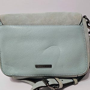 May include: A light blue leather crossbody bag with a suede flap. The bag has a silver-tone chain strap and buckle closure. The brand name is visible on the front. The bag is approximately 9 inches wide and 7 inches tall.