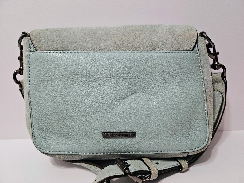 May include: A light blue leather crossbody bag with a suede flap. The bag has a silver-tone chain strap and buckle closure. The brand name is visible on the front. The bag is approximately 9 inches wide and 7 inches tall.