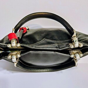 Puede incluir: Bolso de mano de cuero negro con herrajes plateados y un asa curva. El bolso presenta detalles rojos y costuras blancas. El forro interior tiene un estampado colorido. Un accesorio elegante para el uso diario.