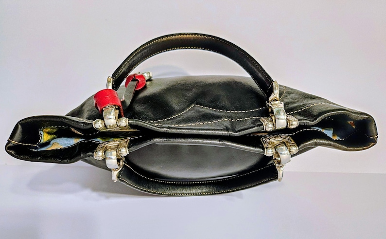 Puede incluir: Bolso de mano de cuero negro con herrajes plateados y un asa curva. El bolso presenta detalles rojos y costuras blancas. El forro interior tiene un estampado colorido. Un accesorio elegante para el uso diario.