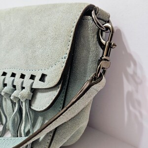 May include: A light blue suede crossbody bag with a flap closure and fringe detailing. The bag features a long strap and silver-tone hardware. The flap has a cut-out design. The bag is made of a soft, textured material.
