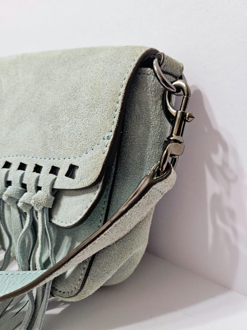 May include: A light blue suede crossbody bag with a flap closure and fringe detailing. The bag features a long strap and silver-tone hardware. The flap has a cut-out design. The bag is made of a soft, textured material.