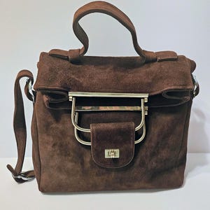May include: A brown suede handbag with a top handle and a detachable shoulder strap. The bag features a rectangular shape with a flap closure and a gold-toned metal clasp. The bag's hardware includes a gold-toned metal bar across the top.