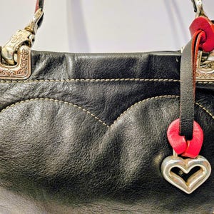 Puede incluir: Bolso de mano de cuero negro con un dise&ntilde;o en forma de coraz&oacute;n cosido en la parte delantera. Cuenta con herrajes plateados, un acento de cuero rojo y un dije de coraz&oacute;n plateado. El bolso tiene una apariencia texturizada y un dise&ntilde;o cl&aacute;sico.