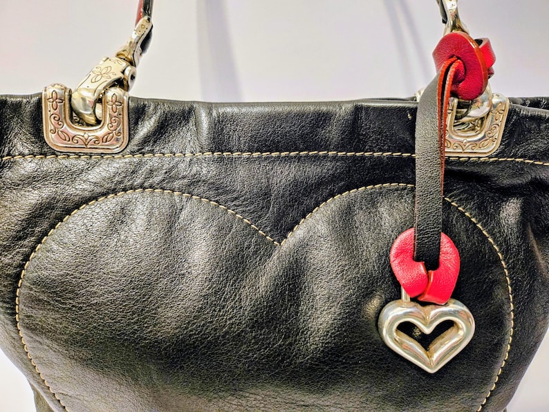 Puede incluir: Bolso de mano de cuero negro con un dise&ntilde;o en forma de coraz&oacute;n cosido en la parte delantera. Cuenta con herrajes plateados, un acento de cuero rojo y un dije de coraz&oacute;n plateado. El bolso tiene una apariencia texturizada y un dise&ntilde;o cl&aacute;sico.