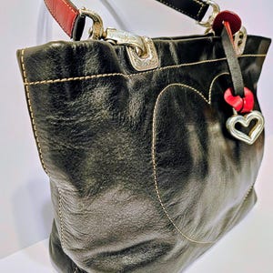 Puede incluir: Bolso tote de cuero negro con un dise&ntilde;o en forma de coraz&oacute;n delineado con costuras color crema. Cuenta con herrajes plateados, un acento rojo en el asa y un dije de coraz&oacute;n. El bolso tiene una forma estructurada y un asa superior.