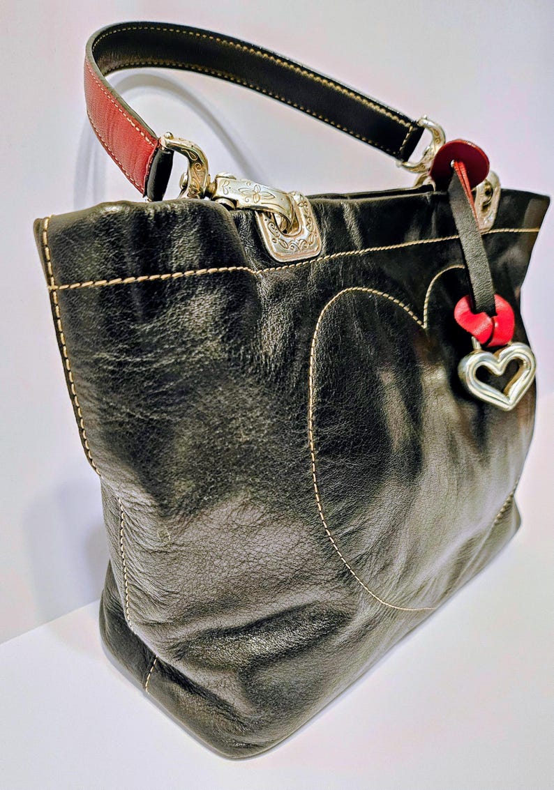 Puede incluir: Bolso tote de cuero negro con un dise&ntilde;o en forma de coraz&oacute;n delineado con costuras color crema. Cuenta con herrajes plateados, un acento rojo en el asa y un dije de coraz&oacute;n. El bolso tiene una forma estructurada y un asa superior.