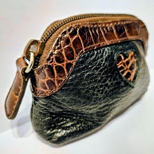 May include: A small, black and brown leather coin purse with a heart-shaped cutout. The top section features a brown, textured, crocodile-like pattern, and a zipper closure. A brown leather pull tab is attached to the zipper.