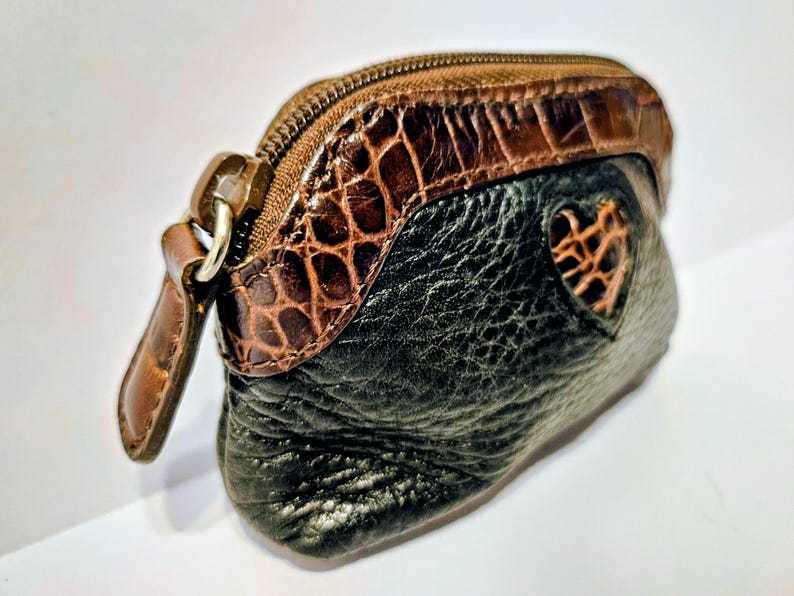 May include: A small, black and brown leather coin purse with a heart-shaped cutout. The top section features a brown, textured, crocodile-like pattern, and a zipper closure. A brown leather pull tab is attached to the zipper.