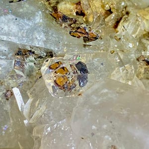 Puede incluir: Primer plano de un cristal de cuarzo transparente con inclusiones de minerales marrones y negros. El cristal está facetado y tiene un brillo vítreo.