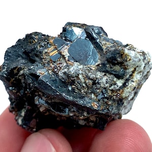 Puede incluir: Primer plano de un espécimen mineral oscuro y multifacético. La roca presenta una mezcla de superficies gris oscuro, negras y plateadas metálicas. Pequeñas inclusiones de color marrón dorado son visibles en toda la piedra.