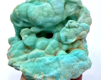 May include: A large, irregularly shaped mineral specimen with a striking turquoise color. The surface has a bubbly, textured appearance with small holes and crevices. The edges have a lighter, more weathered look. The mineral is held by a hand.