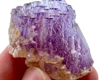 May include: A natural fluorite mineral specimen. The crystal features a layered structure with shades of purple and white. The specimen has a rough, textured surface and is held in a hand.